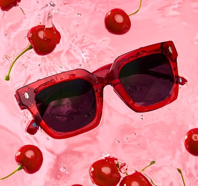 Zenni square-, red sunglasses with dark tinted lenses are shown in water with cherries floating around them.